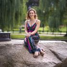 A picture of Jessica Bloch in a sun dress sitting outside on a large stone with the base of a lamp post beside her. There are trees all around and it is sunny.