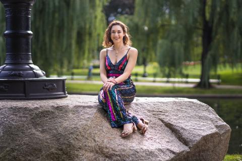 A picture of Jessica Bloch in a sun dress sitting outside on a large stone with the base of a lamp post beside her. There are trees all around and it is sunny.